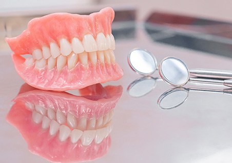 Complete set of dentures next to dental mirrors on reflective surface